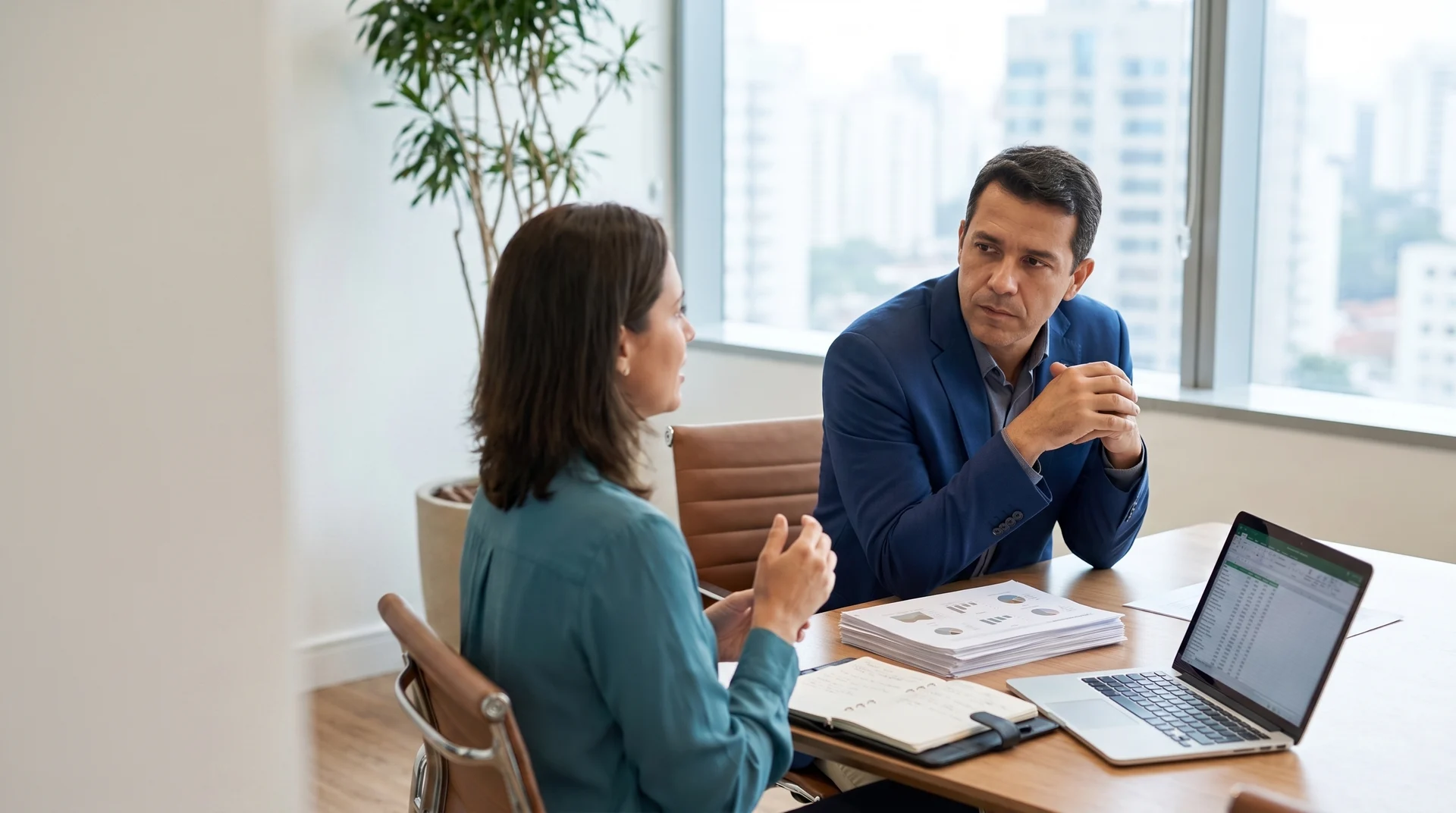 Duas pessoas em uma reunião de trabalho, com relatórios e notebook sobre a mesa.